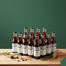 Load image into Gallery viewer, Flat lay photography of twelve bottles of deep red Sorrel hibiscus drink elegantly arranged on a dark green background with festive Christmas decorations, including gold ribbons, small white gift boxes, and evergreen pine branches.