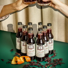 Load image into Gallery viewer, A flat lay of Brooklyn Brewed Sorrel, featuring a large bottle and a small bottle of the healthy, nut-free spiced hibiscus roselle mocktail. Two hands toast with glasses of the deep red, alcohol-free beverage over a green table scattered with dried hibiscus petals and orange peel. This premium, Caribbean-inspired, and Black-owned non-alcoholic drink is perfect for Dry January, Passover, and holiday gifting.